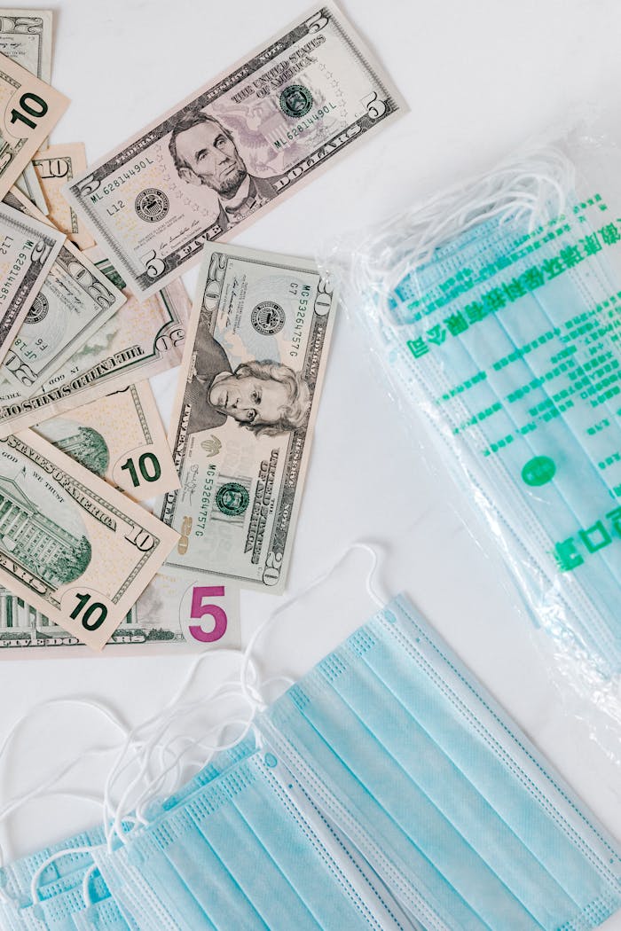 services-02 Top view composition of disposable medical masks placed on white table near scattered randomly various paper banknotes of American dollars