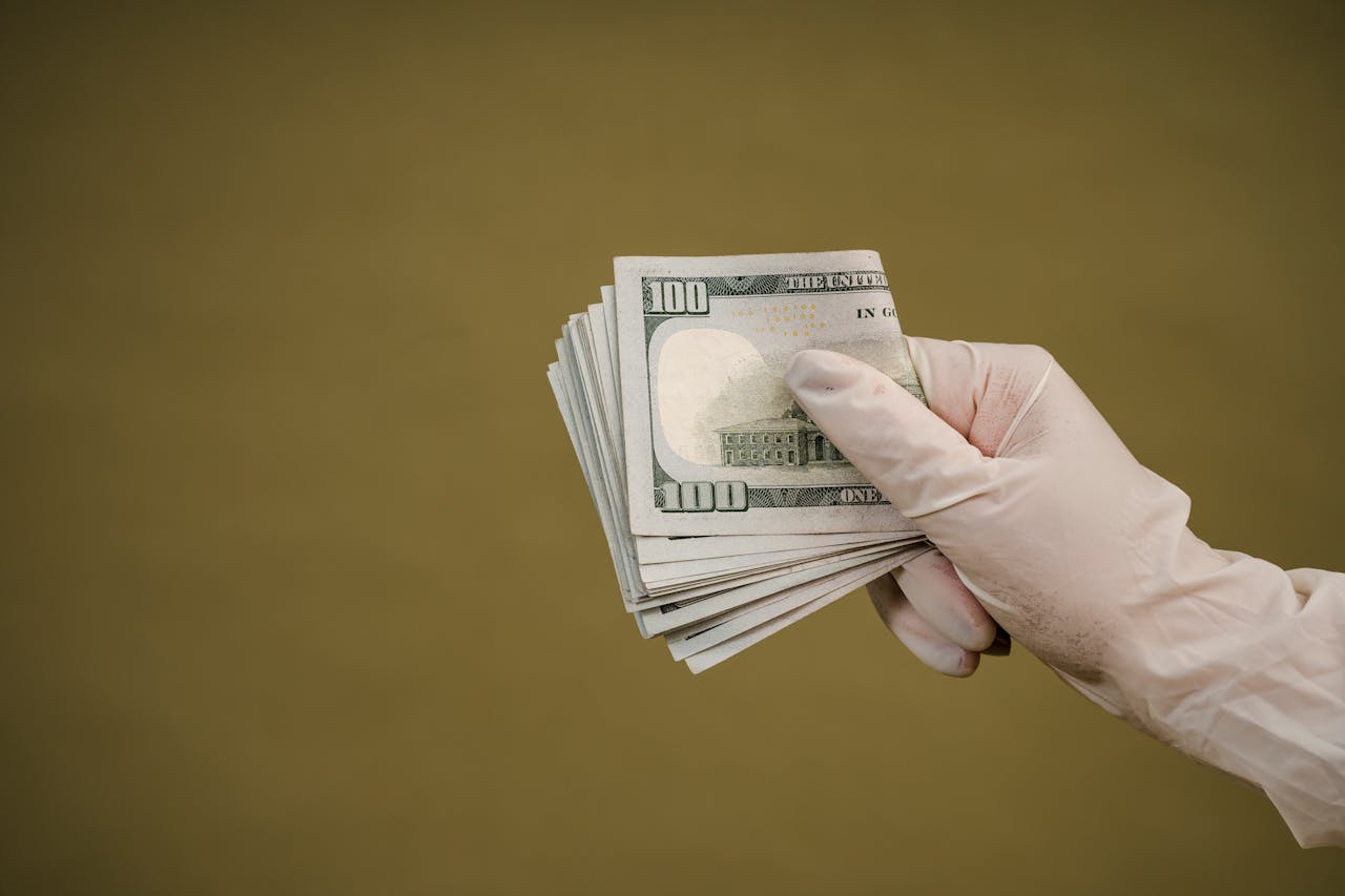 home-hero A gloved hand holds a stack of hundred-dollar bills against a plain background.
